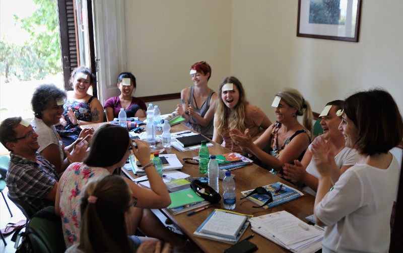 A group of people sit around a table with books and bottles, laughing and clapping. Some have sticky notes on their foreheads. The setting appears to be a casual indoor meeting or workshop.