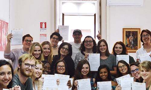 A group of people stands indoors, each holding a certificate, smiling at the camera. There is an open window behind them, and art on the wall.