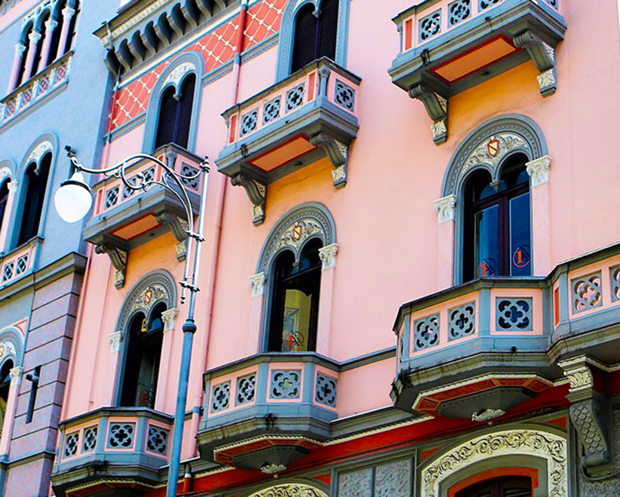 Colorful building facade with intricately designed windows, small decorative balconies, and an ornate street lamp in front. The walls are painted in pink and gray hues.