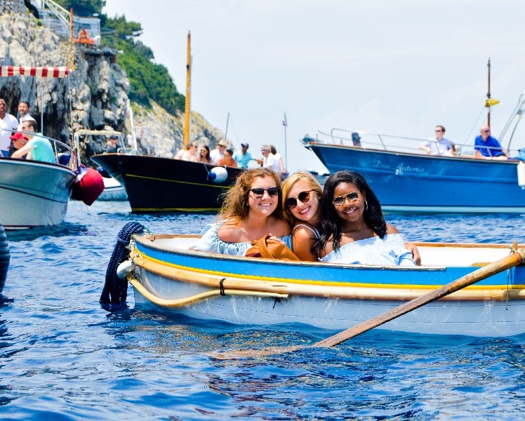 Three women are smiling and sitting in a small boat on a sunny day, surrounded by other boats on a body of water. Cliffside can be seen in the background.