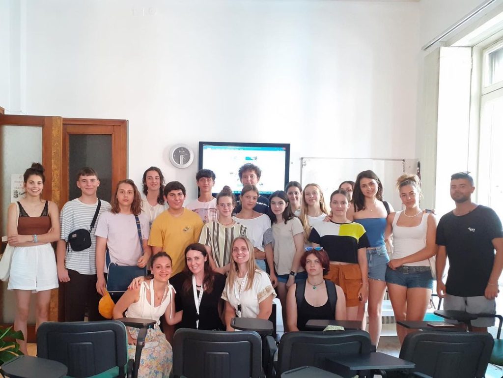 A group of 20 people, mostly young adults, pose together in a well-lit classroom. They stand in front of a large screen with "Salerno" and "Napoli" written on it, wearing casual summer clothes, and most are smiling at the camera.
