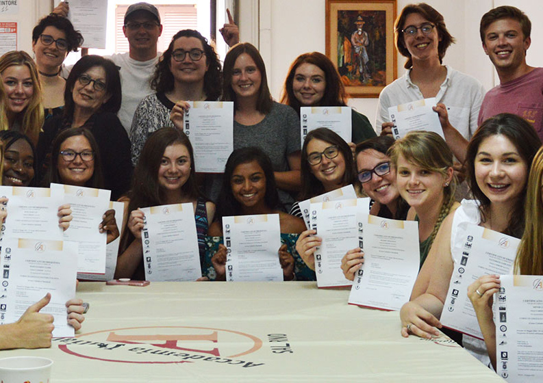 Un gruppo di persone sorridenti con in mano dei certificati posa per una foto attorno a un tavolo coperto da un panno bianco.