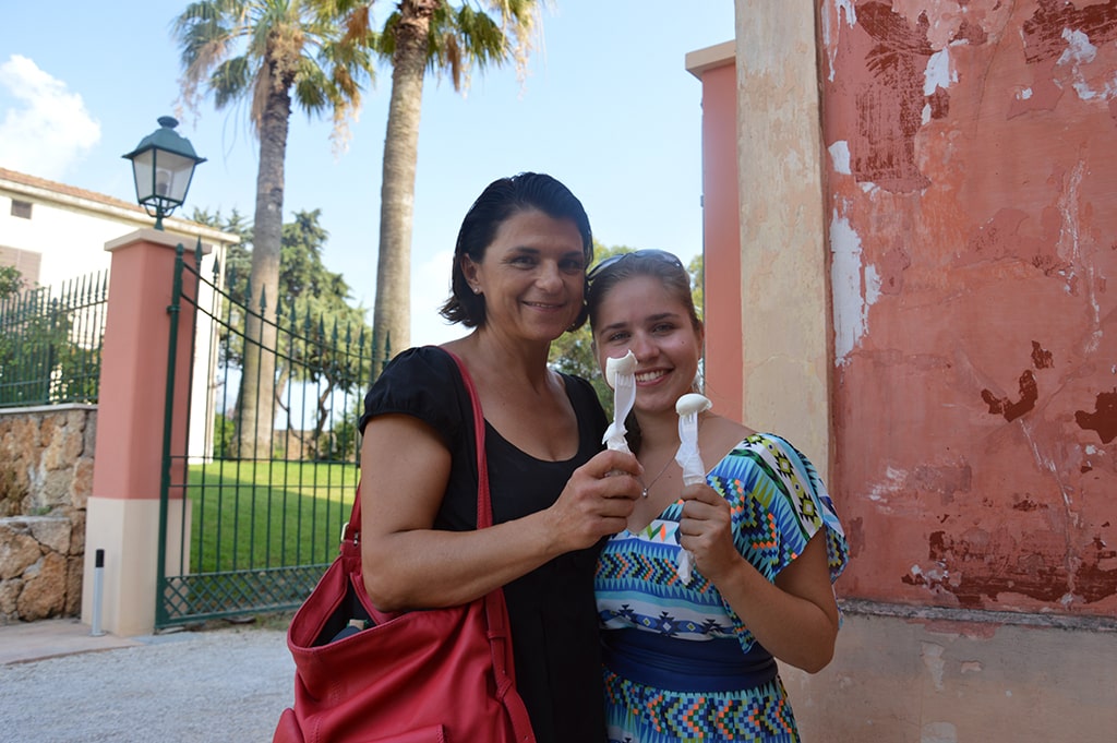 Two women are standing outdoors in front of a gate and a peach-colored wall, each holding white ceramic objects resembling mushrooms. The woman on the left has a red bag. Palm trees are in the background.