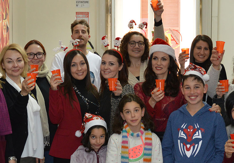 A group of people, including children, wearing festive hats and holding orange cups, smile at the camera in a well-lit room at Accademia Italiana Salerno.