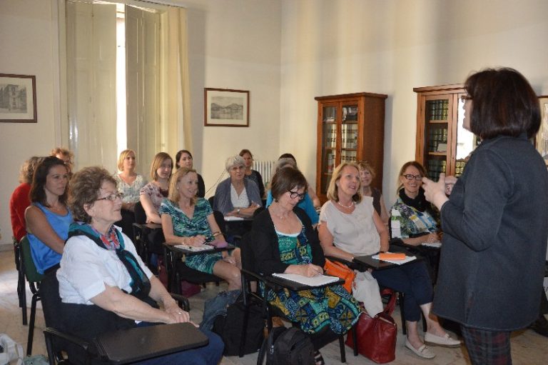 Un gruppo di donne siede attento in un'aula a Salerno mentre una donna in prima fila parla loro. Alcuni prendono appunti, catturando ogni dettaglio come se fossero a Napoli.