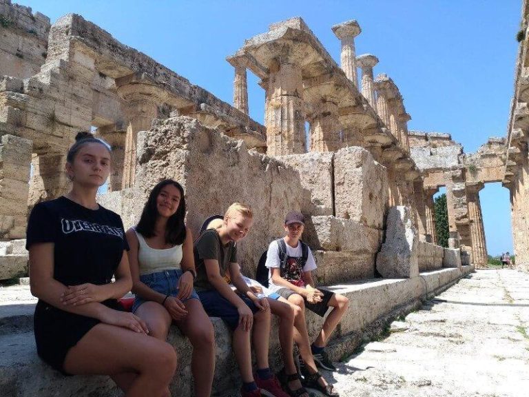 A group of four young people sit and stand among the ruins of an ancient stone structure on a sunny day.