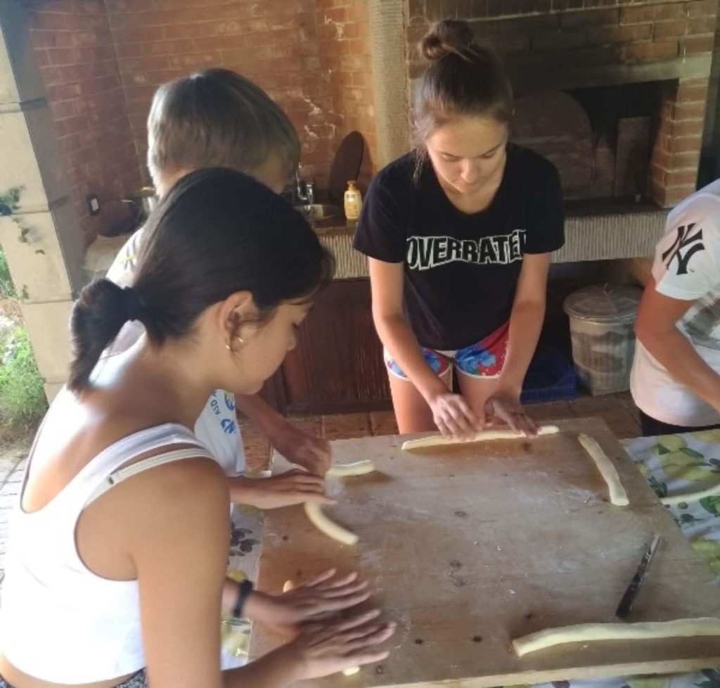 Four people are standing around a table, rolling dough into long strands. There is a brick oven in the background.