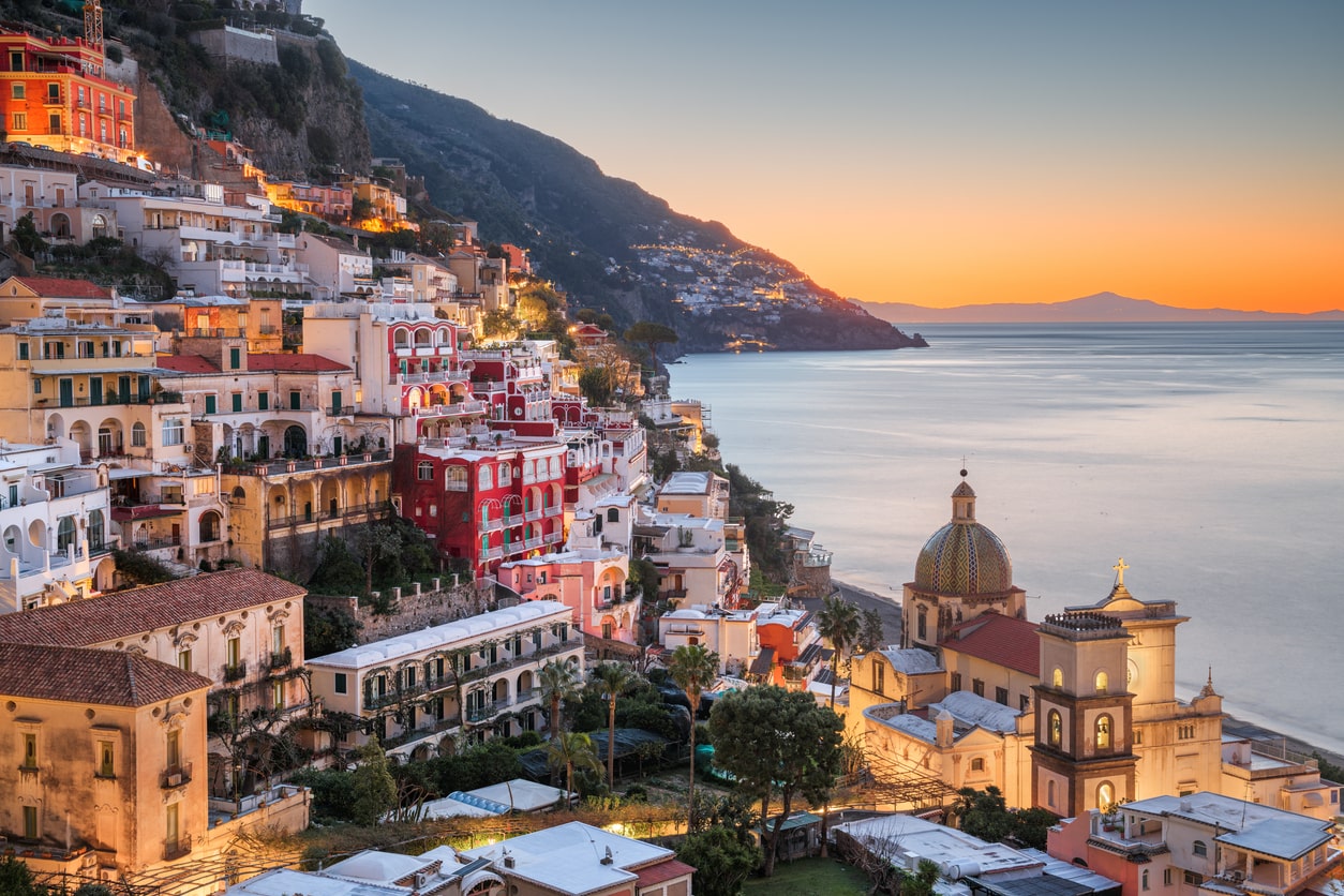 Colorful buildings on a hillside overlook a calm sea at sunset in Positano, Italy, with a prominent domed church in the foreground